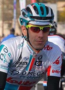 A man in his mid twenties wearing a cycling jersey that is white, red, and two tones of blue. He also wears sunglasses with yellow frames, and another cyclist is vaguely visible behind him.