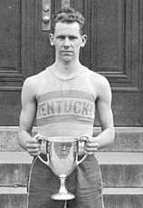 A young man in a sleeveless shirt with the word "Kentucky" on it holds a cup-like trophy in front of his stomach and stares intently at the camera