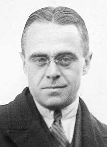 Black and white photo of a bespectacled man in suit and tie looking at the camera