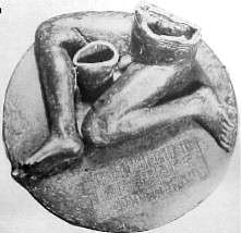 Black-and-white photograph of a statue consisting of an inscribed, round pedestal on top of which sits a seated, nude, male figure of which only the legs and lower torso are preserved