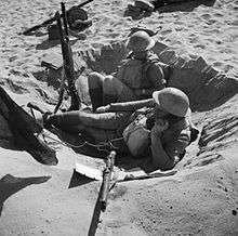 A black and white photograph of two men wearing military uniforms and helmets in a foxhole in the desert; one man operates a field telephone