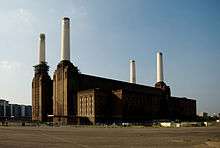 Colour picture of a power station factory with four tall white chimneys. The image was taken on a sunny day. The sky is blue and the building is brown.