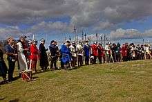 A line of people in mediaeval dress and armour, several with weapons, stand under a cloudy sky.