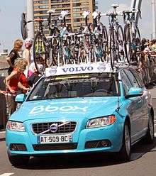 A powder blue automobile with several bicycles attached to its roof. Spectators at the roadside are looking behind the car to something out of the frame.