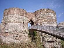 Two round towers of light yellow stone at the bottom and dark orangy stone at the top on either side of an arched entrance. A bridge leads from the entrance to allow access.
