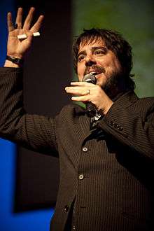 Brown-haired man wearing a dark brown shirt holding a microphone and waving