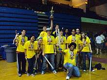 Twelve adolescents pose for a picture, standing and smiling. They are holding foam swords, a model of a human body's muscular system, a trophy, and medals.