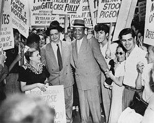 Two men dressed in suits are surrounded by persons holding signs.
