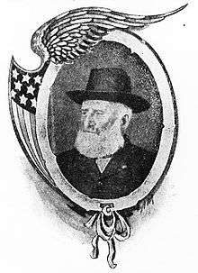 Head and shoulders of a white man with a bushy white beard, wearing a dark suit coat and wide-brimmed hat. The portrait is in a decorative frame featuring a bird's wing and stars and stripes.