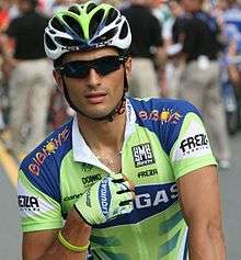 A cyclist wearing a green and blue jersey with white trim, a matching helmet and glove on his right hand (the only one visible), and sunglasses. His right hand hovers around the zipper on the front of his jersey, and vaguely visible behind him are three people in black shirts and khaki pants.