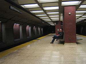 A near-empty station platform of an underground train station, with a man sitting on a bench