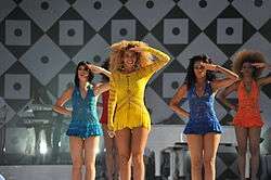 A woman in a yellow dress, flanked by three female dancers, salutes to the crowd