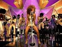 A female is singing and dancing. She wears a golden dress and heels of the same color. Behind her, a band is playing their instruments.