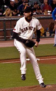 A man in a white baseball uniform with the word "GIANTS" written across it prepares to throw a baseball with his left hand to home plate during a game.