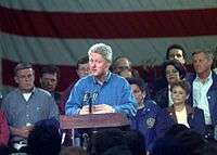 Bill Clinton is standing at a podium speaking to a crowd. The former mayor of Grand Forks is at the right of the image.