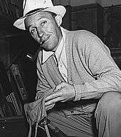 A man wearing a grey sweater and a fedora squats near an open box, holding a small canvas bag.