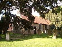 A stone church with a red tiled roof seen between trees in a churchyard.  On the west gable is a bellcote.