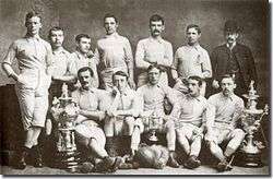 A group of men, most of whom are wearing shorts and football shirts, holding two trophies, one of which is the FA Cup