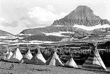 A black-and-white photograph.  Against a backdrop of mountains and forests is a row of eight tipis.