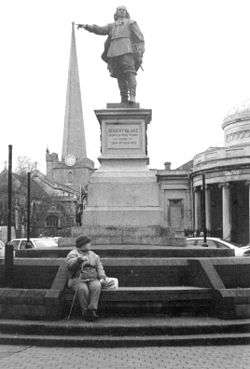 In the foreground is a statue of a man on a plinth above steps, with person sitting on them. In the background is a church tower. The picture is arranged so that the outstretched arm with a pointing finger of the figure appears to be touching the top of the tower.
