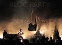 Two guitarists, a drummer, and a bassist are performing on a smoky stage lit by white stage lights. The stage background is a black tarpaulin emblazoned with "BLOC PARTY." and a large black-and-white cityscape below it. Most of the crowd have their hands up in the foreground, which also includes a waving flag.