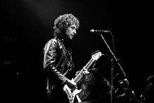 Black and white, waist high portrait of middle aged man with curly hair, leather jacket, electric guitar.