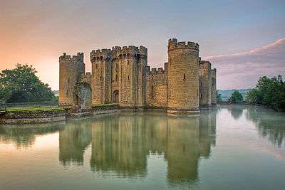 Photo of Bodiam Castle at sunset with towers and battlements reflected in a wide moat. A path leads away from the entrance of the castle across two islands.