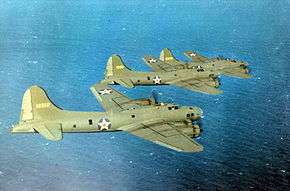 Three olive green four-engined propeller aircraft fly over the ocean.