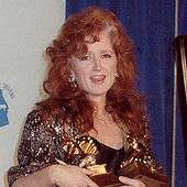 A woman with curly red hair wearing a sparkling jacket and holding three gold trophies.