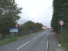 A photograph facing west along Bothkennar Road with a sign showing the entrance to the village of Carronshore.