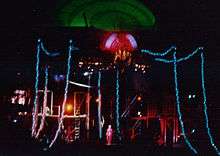 An image of a stage with a giant luminescent spider overhead, its body glowing green, its head glowing red and its legs glowing blue. Below the spider, tiny human forms can be see on stage