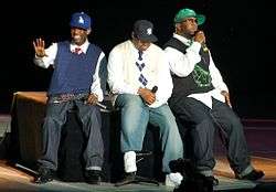 Three African-American men sitting next to each other on a black stage. They are all wearing caps with jeans and sneakers.