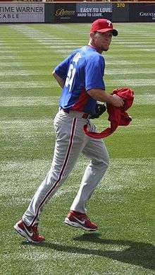 A man in grey pants, a blue baseball jersey, and a red baseball cap with "P" on it jogs in the field.