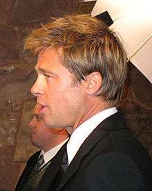 A side view of a man, who is facing to the left, with light brown hair. He is wearing a black suit and tie with a white shirt. Another male, also wearing a suit, is visible in the background.