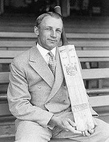 Man in double breasted suit, hair parted down the middle, sitting on a long bench in a sports stadium, posing with a cricket bat, held vertical and supported on his thigh.