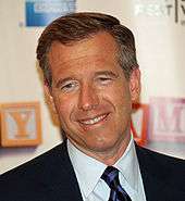 A head shot of Brian Williams, a caucasian male in his late-40s with dark hair. He smiles, and wears a black suit with a white shirt and purple and black-striped tie.