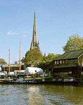 A tall church spire over a quayside with wooden sheds and boats covered with tarpaulins. In front of these on the water a twin masted sailing boat and a narrowboat