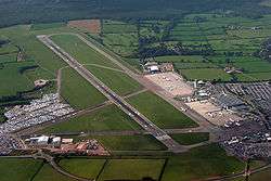 An aerial view of an airport with one main runway, car parks on the left and right, and aircraft parked outside terminal buildings on the right.