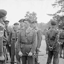 General officer with gloves and cane in hand, talking to a sergeant, surrounded by other officers and men