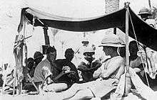 A number of men dressed in shorts and shirts with sleeves rolled up, one in a singlet sit in the shade of an awning. Several wear pith helmets, one of whom sits in the sun shirtless outside the awning leaning against a pole holding up the awning.