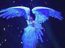 A female blond performer. She is wearing a white winged dress and sings as she's lifted in the air.