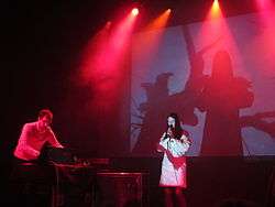 An electronic music band performing onstage. To the left a man is performing on a synthesiser; to the right a woman is singing with a microphone, with her shadow visible on a projected background behind her. Pink and purple stage lights are visible.