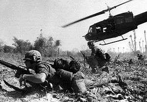 A black and white image of a man crawling across the ground with a helicopter flying in the background