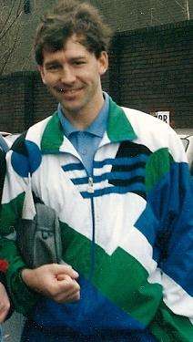 The upper body of a brown haired man. He has a grey bag under his arm, is wearing a blue buttoned-up shirt underneath a white, black, green and blue tracksuit top.