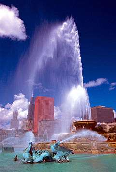 A fountain spouting water from several points with several tall buildings and a blue sky in the background.