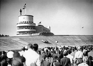 A large crowd of people watch a streamlined single-seater drive around a massive curved racing bank. A Nazi flag is flying in the distance atop a tower.