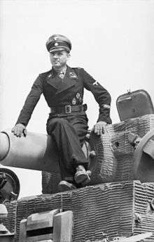 Tank Ace Michael Wittmann, wearing Waffen SS dress uniform, sits atop the main gun of his Tiger tank. The tank is covered in a ridged paste.