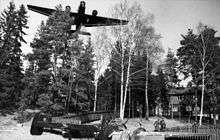 A black-and-white photo of a three-engine aircraft flying over trees. The aircraft is viewed from the front and below. Among the trees is a house with three people standing in front of it. A further aircraft is sitting on the ground and viewed from the rear-right.