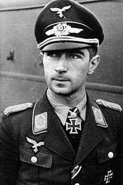 The head and shoulders of a young man, shown in semi-profile. He wears a field cap and a pilot's leather jacket with a fur collar, with an Iron Cross displayed at the front of his shirt collar. His hair is dark and short, his nose is long and straight, and his facial expression is a determined and confident smile; his eyes gaze into the distance.