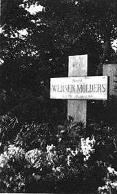 Black-and-white photograph of a wooden cross on a grave, bearing the inscription "Oberst Werner Mölders, 18. 3. 1913 – 22. 11. 1944." The name Werner Mölders is in large letters. Trees are seen in the background; the area in front of the cross is covered with low-growing plants bearing flowers.